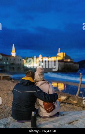 Un homme et une femme s'assoient sur le bord de la mer et regardent les vieilles maisons illuminées. Vue arrière Banque D'Images