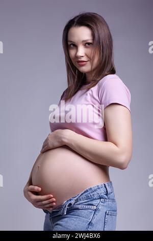 Jeune femme enceinte souriante en t-shirt rose et jeans sur fond gris. Banque D'Images