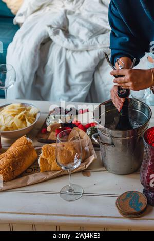 femme en chemise bleue ouvrant une bouteille de vin frais dans un seau à champagne sur une table basse avec des collations Banque D'Images