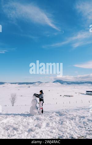 Maman et petite fille font un bonhomme de neige, debout sur une clairière enneigée dans une vallée de montagne. Vue arrière Banque D'Images