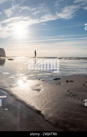 Cette image évocatrice capture une figure solitaire enveloppée dans la douce étreinte des rayons du soleil sur une plage tranquille. Le soleil, placé juste au-dessus de l'horizon, baigne la scène dans une lumière douce et diffuse, créant un sentiment de paix et d'immobilité. Le sable humide autour de la figure reflète la lumière du soleil, ajoutant une qualité chatoyante à la scène, tandis que les empreintes menant à la figure suggèrent un voyage pris dans la solitude. Le ciel croustillant, parsemé de nuages délicats, encadre ce moment de réflexion tranquille. Figure solitaire dans l'étreinte du Soleil. Photo de haute qualité Banque D'Images