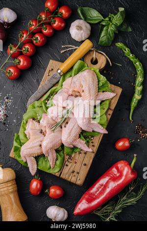 Ailes de poulet crues avec des ingrédients pour la cuisson sur une planche à découper en bois sur fond de pierre sombre Banque D'Images