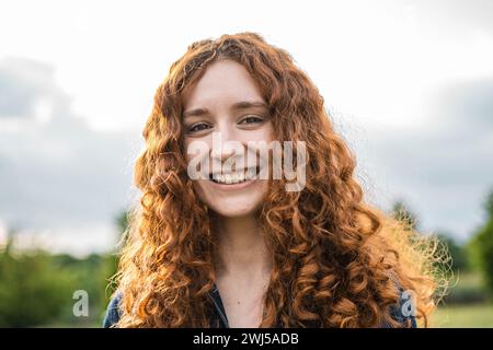 Femme rousse avec des cheveux bouclés souriant dans la lumière extérieure naturelle - Un portrait capturant la joie jeune et la beauté naturelle. Banque D'Images