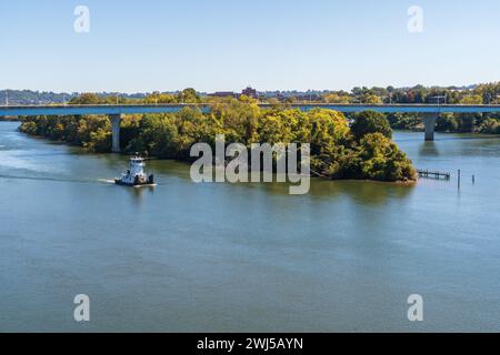 Un petit remorqueur blanc passe devant une île couverte d'arbres dans la rivière Tennessee sous un long pont autoroutier à Chattanooga, dans le Tennessee. Banque D'Images