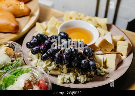 Une variété de fromages gastronomiques accompagnés de raisins noirs frais et d'un bol de miel, parfait pour une configuration de planche à fromages ou un événement d'appariement de vin. Banque D'Images