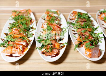 Assiettes blanches remplies de petits pains de saumon fumé recouvertes de roquette fraîche, de fromage à la crème et arrosées de glaçage balsamique, prêtes à servir. Banque D'Images