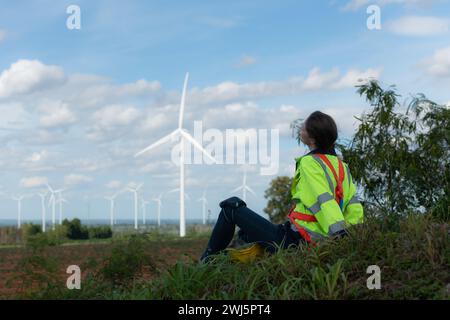 Ingénieur féminin assis se détendre sur l'herbe avec des éoliennes en arrière-plan. Banque D'Images