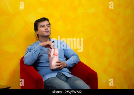 Jeune homme mangeant du pop-corn assis dans un fauteuil rouge sur un fond jaune attendant de regarder un film. Banque D'Images