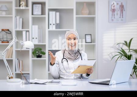 Une médecin musulmane professionnelle portant un hijab rayonne de positivité alors qu'elle consulte des patients dans son bureau lumineux de clinique. Banque D'Images