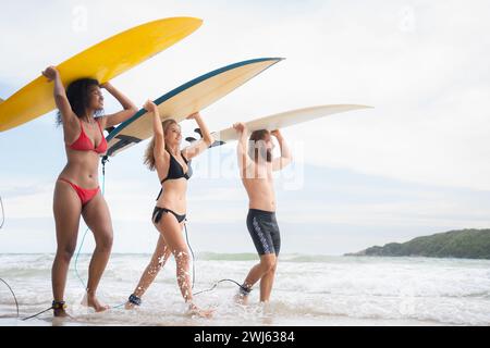 Vue arrière de deux femmes et d'un jeune homme tenant des planches de surf sur leur tête. et marcher dans la mer pour surfer Banque D'Images