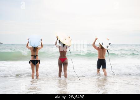 Vue arrière de deux femmes et jeune homme tenant des planches de surf sur leurs têtes et marchant dans la mer pour surfer Banque D'Images