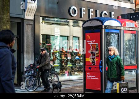Kingston-upon-Thames, Londres, Royaume-Uni, 12 février 2024, femme portant un sac à provisions et homme sur vélo passant des boîtes téléphoniques publiques et un magasin de chaussures de bureau Banque D'Images