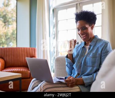 Femme excitée à la maison avec ordinateur portable en utilisant la carte de crédit pour faire l'achat en ligne ou réserver des billets Banque D'Images