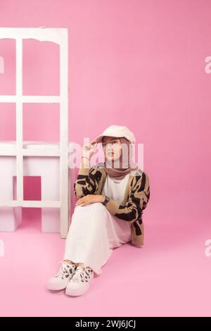 Portrait d'une belle jeune femme assise les jambes croisées sur un sol rose isolé avec des lunettes et un chapeau dans un déshérité blanc et un pull marron, coréen Banque D'Images