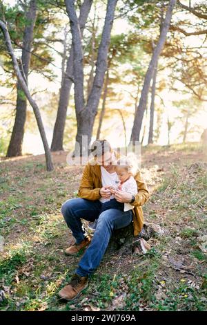 La petite fille est assise sur les genoux de son père dans la forêt et joue avec des cailloux dans sa main Banque D'Images