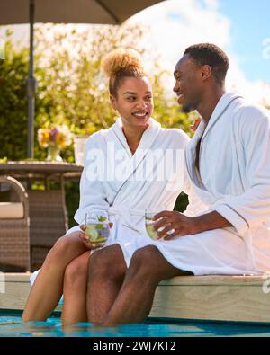Couple portant des peignoirs à l'extérieur assis avec des boissons aux pieds dans la piscine le jour du spa Banque D'Images