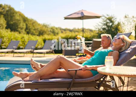 Couple senior en vacances avec femme portant des écouteurs sans fil et homme lisant livre par piscine Banque D'Images