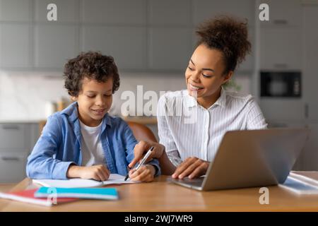 Mère noire aide son fils avec les devoirs sur ordinateur portable dans la cuisine bien éclairée Banque D'Images