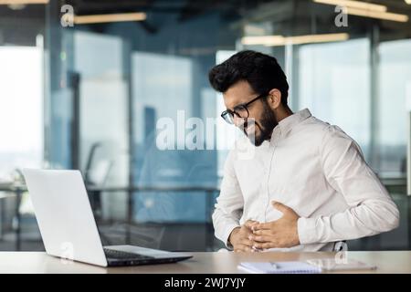 Un jeune homme d'affaires indien souffre de graves douleurs à l'estomac. Assis dans le bureau au bureau et se tenant les mains, plié et grimaçant dans l'inconfort. Banque D'Images