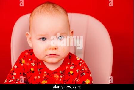 Portrait d'un enfant mignon dans des vêtements rouges avec un visage sale après avoir mangé, assis sur une chaise haute sur un fond rouge Banque D'Images