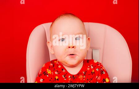 Portrait d'un enfant mignon dans des vêtements rouges avec un visage sale après avoir mangé, assis sur une chaise haute sur un fond rouge Banque D'Images