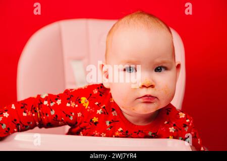 Portrait d'un enfant mignon dans des vêtements rouges avec un visage sale après avoir mangé, assis sur une chaise haute sur un fond rouge Banque D'Images