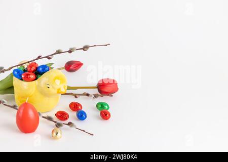 Oeufs en chocolat colorés dans un poussin en céramique jaune à côté de chatons de palmier, tulipes et bougie, fond blanc, salle de copie Banque D'Images