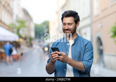 Un jeune homme souriant se tient debout dans une rue de la ville dans une chemise en Jean et utilise un téléphone portable. Gros plan. Banque D'Images