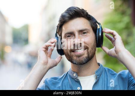 Un homme mûr heureux avec une barbe marche à travers la ville au crépuscule, appréciant ses chansons préférées sur ses écouteurs. Banque D'Images