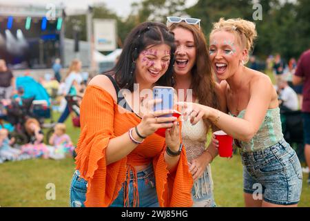 Trois amies féminines portant des paillettes regardant le téléphone portable au Festival de musique d'été tenant des boissons Banque D'Images