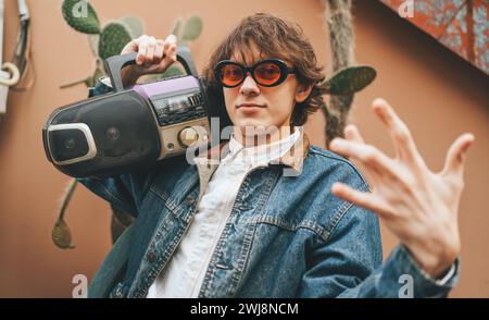 Jeune homme élégant hipster porter dans la veste en denim rétro de style ancien des années 90 tenir lecteur de cassette de bande audio et montrer des gestes par les doigts comme chanteur de rap. Banque D'Images