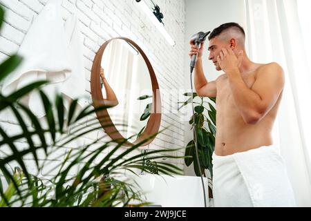 Jeune homme attrayant séchant ses cheveux après la douche dans la salle de bain Banque D'Images