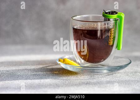 Une tasse transparente de thé chaud avec une tranche de citron sur une soucoupe et un infuseur de thé vert suspendu sur le côté Banque D'Images