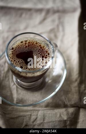 Gros plan détaillé capturant la texture mousseuse d'un café fraîchement préparé dans une tasse en verre transparent sur fond de tissu texturé Banque D'Images