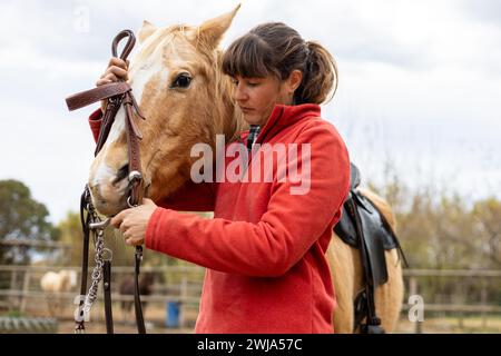 Jeune cavalière mettant les rênes et la bride sur son cheval brun dans un centre équestre Banque D'Images