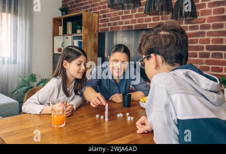 Une mère avec ses enfants souriant et jouant à un jeu de dés à la table familiale avec des collations et des boissons pendant un week-end confortable à la maison Banque D'Images