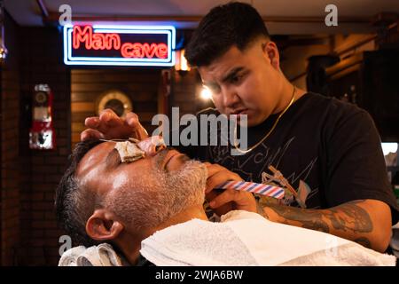 Vue latérale de l'homme barbu se relaxant sur une chaise avec protecteur d'oeil et serviettes rond cou tout en obtenant la barbe rasée par le barbier dans le magasin contre le néon signe sur Banque D'Images