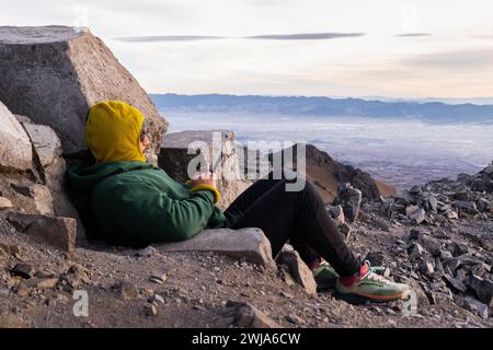 Vue latérale complète du corps du voyageur masculin méconnaissable dans la capuche et veste chaude avec pantalon et bottes assis sur la falaise rocheuse et en utilisant un smartphone au Nevado Banque D'Images