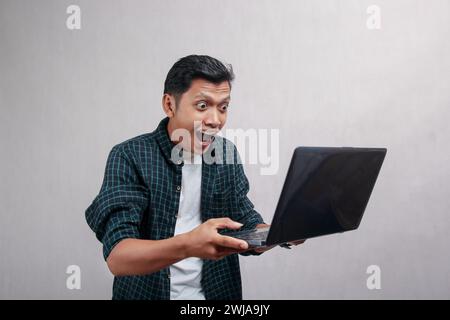 Un homme dans une chemise vert Vichy tient un ordinateur portable avec une main levée et une expression surprise. Banque D'Images