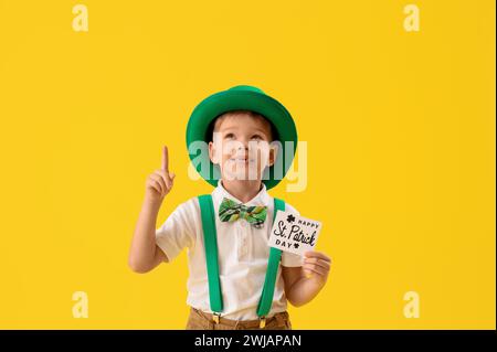 Petit garçon mignon avec le chapeau de leprechaun et la carte de voeux pointant vers quelque chose sur fond jaune. Prog Célébration de la fête de Patrick Banque D'Images