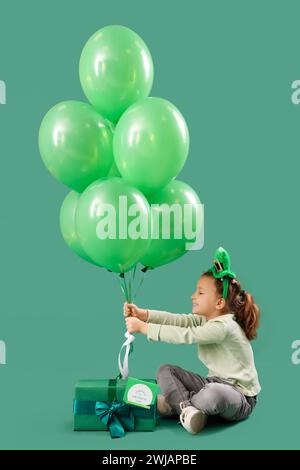 Petite fille mignonne avec chapeau de leprechaun, boîte cadeau et ballons sur fond vert. Prog Célébration de la fête de Patrick Banque D'Images