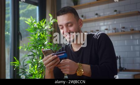 Homme souriant tenant la carte de crédit et smartphone saisit le numéro de carte de crédit pour les achats en ligne. Masculin utilisant des paiements mobiles instantanés faciles, faisant l'achat Banque D'Images