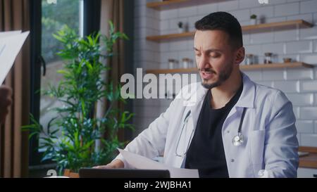 Médecin expérimenté portant un manteau blanc prenant des rapports de patient de son assistant et des examens. Praticien masculin faisant de la paperasse Banque D'Images