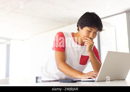 Adolescent asiatique étudie attentivement sur son ordinateur portable dans un bureau lumineux Banque D'Images