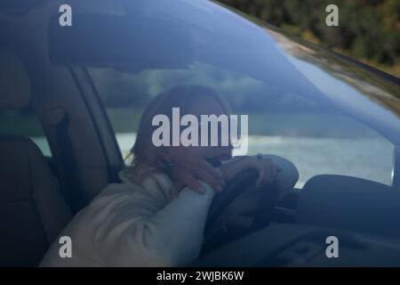 Une femme blonde dans un pull blanc et un Jean conduit. Femme heureuse assise dans une voiture avec un intérieur blanc. Banque D'Images