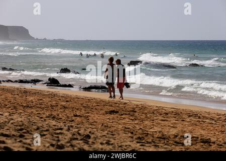 Couple d'âge mûr profiter d'une promenade se tenant la main le long d'une plage de sable, regarder les surfeurs, Piedra Playa, El Cotillo, Fuerteventura prise novembre 2023 Banque D'Images