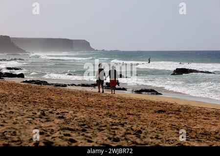 Couple d'âge mûr profiter d'une promenade se tenant la main le long d'une plage de sable, regarder les surfeurs, Piedra Playa, El Cotillo, Fuerteventura prise novembre 2023 Banque D'Images