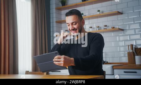 Homme ravi jouant à un jeu vidéo sur une tablette tout en étant assis dans la cuisine à la maison. Homme concentré excité engrossé dans la fantaisie de course intéressante Banque D'Images