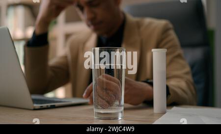 Surtravaillé homme d'affaires afro-américain malade jeter comprimé analgésique dans un verre d'eau appliquer pilule pétillante avec des bulles dans la boisson sur la table de bureau souffrir Banque D'Images