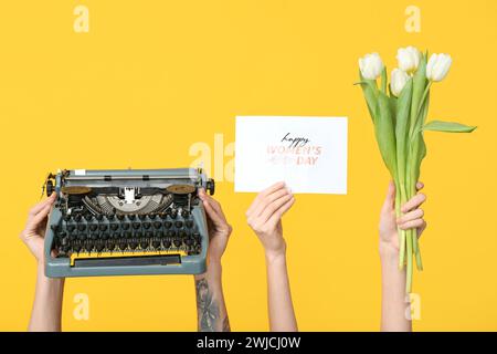 Mains tenant une machine à écrire vintage, des tulipes et une carte postale festive avec texte HAPPY WOMEN'S DAY sur fond jaune Banque D'Images